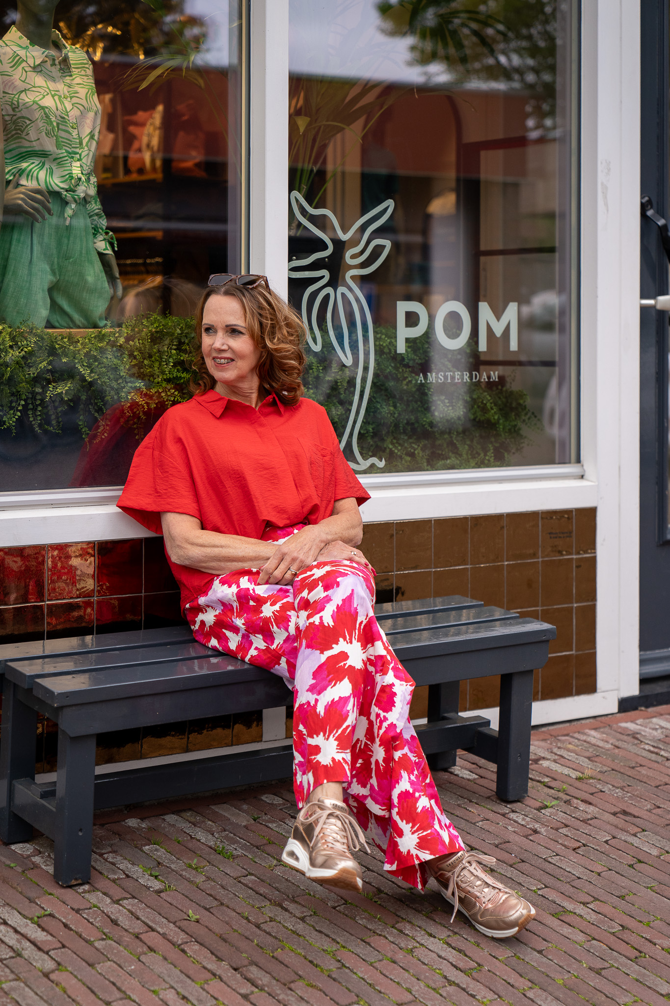 Sytha Sr zit op bankje voor winkel in Winsum in een rode blouse van POM